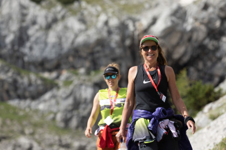 Zwei Frauen in sportlicher Kleidung, mit Sonnenbrille und Schlüsselband wandern auf einem felsigen Bergpfad im Freien. Beide lächeln, wobei die vordere Frau eine Mütze und eine um die Taille gebundene Jacke trägt.