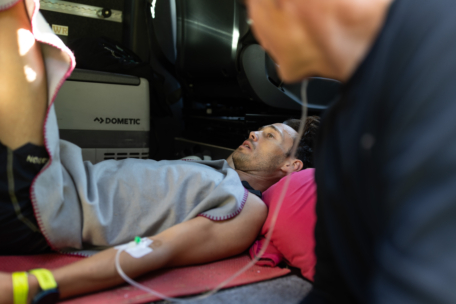Ein Mann liegt auf dem Rücken auf einer Matte mit einer Infusion im Arm, bedeckt mit einer Decke, und schaut nach oben. Eine weitere Person befindet sich unscharf im Vordergrund. Die Szene scheint sich in einem Fahrzeug oder einem geschlossenen Raum zu befinden.