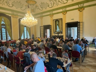 Eine große Gruppe von Menschen, darunter Kinder und Erwachsene, spielt Schach in einem großen, verzierten Raum mit Kronleuchtern, hohen Decken und verzierten Wänden. Das Sonnenlicht strömt durch hohe Fenster mit grünen Vorhängen herein.