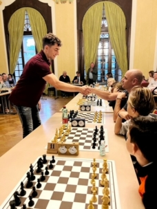 Ein junger Mann schüttelt seinem Gegner in einem großen Raum, umgeben von anderen Spielern und Zuschauern, die Hand und bereitet sich auf ein Schachspiel vor.