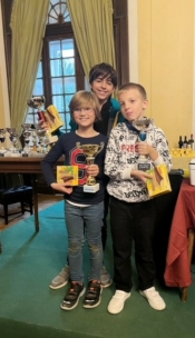 Drei lächelnde Kinder stehen dicht beieinander in einem Raum und halten Trophäen und verpackte Preise in der Hand. Hinter ihnen befindet sich ein Tisch mit weiteren Trophäen und ein großes Fenster mit grünen Vorhängen, die das Tageslicht hereinlassen.