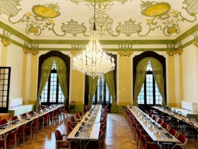 Ein großer, eleganter Raum mit verzierten Decken, hohen Fenstern mit grünen Vorhängen, einem großen Kronleuchter und langen Tischen mit Schachbrettern und roten Stühlen, bereit für ein Schachturnier.