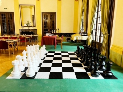 In einem eleganten Raum mit hohen Fenstern, Holztüren und Tischen, die im Hintergrund für ein Schachturnier aufgestellt sind, steht ein großes Schachspiel mit übergroßen schwarzen und weißen Figuren auf einem karierten Matt.