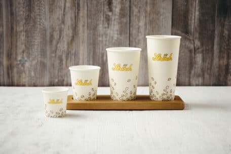 Vier unterschiedlich große Pappbecher mit Mr. Shock und einem Kaffeebohnenmuster stehen in einer Reihe auf einem Holztablett vor einem hölzernen Hintergrund.