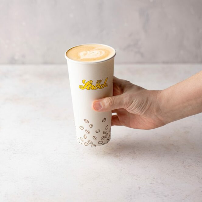 Eine Hand, die einen großen weißen Pappbecher mit Latte Art auf der Oberseite hält. Der Becher hat ein gelbes Shocks-Logo und ein Muster von Kaffeebohnen in der Nähe des Bodens. Der Hintergrund ist eine helle, neutrale Fläche.