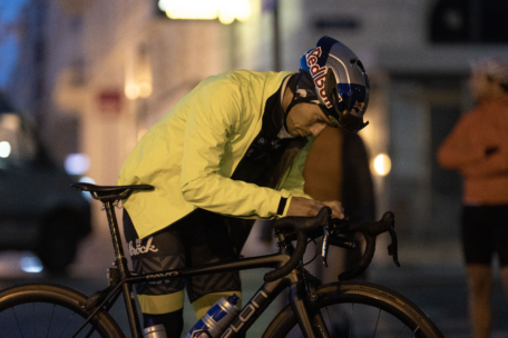 Ein Radfahrer mit gelber Jacke und Red-Bull-Helm beugt sich über den Lenker eines schwarzen Fahrrads und stellt etwas ein, während eine andere Person im Hintergrund verschwommen zu sehen ist.