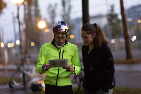 Zwei Personen stehen im Freien neben Fahrrädern. Einer trägt einen reflektierenden Helm und eine hellgrüne Jacke und schaut auf eine Karte. Die andere, dunkel gekleidet, lächelt neben ihnen. Es scheint früher Morgen oder Abend zu sein, die Straßenlaternen sind eingeschaltet.