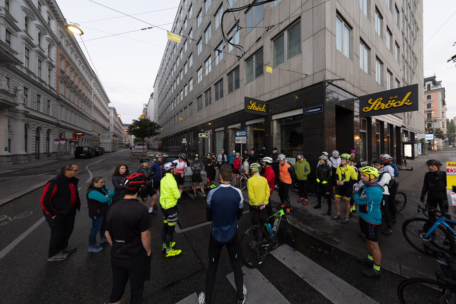 Eine Gruppe von Radfahrern mit Helmen und Sportkleidung versammelt sich an einer Straßenecke in der Nähe einer Bäckerei namens Ströck und bereitet sich auf eine Fahrt in der Morgendämmerung vor.