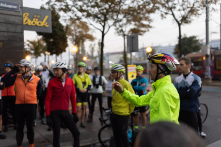 Eine Gruppe von Radfahrern mit Helmen und leuchtenden Jacken versammelt sich frühmorgens auf einer Stadtstraße und hört einer Person in einer neongelben Jacke zu, die spricht und mit der Hand gestikuliert.