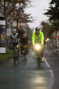 Eine Gruppe von Radfahrern fährt in der Abenddämmerung auf einem städtischen Radweg. Der führende Radfahrer trägt eine leuchtend gelbe Jacke und einen Helm mit eingeschaltetem Frontlicht, während die anderen in Radfahrerkleidung hinterherfahren. Der Weg ist von Bäumen und Gebäuden gesäumt.