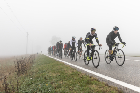 Eine Gruppe von Radfahrern fährt in einer Reihe auf einer Landstraße durch dichten Nebel, mit Feldern auf beiden Seiten und schlechter Sicht im Hintergrund.