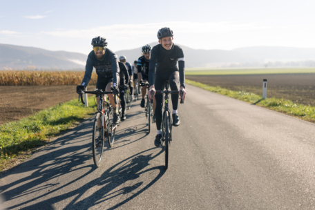Eine Gruppe von Radfahrern, die Helme und Fahrradkleidung tragen, fährt gemeinsam auf einer Landstraße, die von Feldern umgeben ist, mit Hügeln im Hintergrund unter einem klaren Himmel.