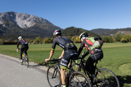 Drei Radfahrer fahren auf einer Landstraße, umgeben von grünen Feldern und Bäumen, mit einem Berg und blauem Himmel im Hintergrund. Die Radfahrer tragen Helme und sportliche Fahrradkleidung.