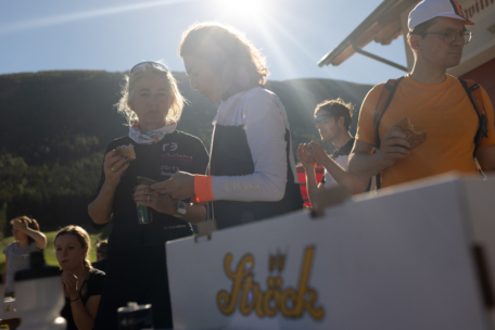 Mehrere Menschen stehen im Freien im Sonnenlicht, essen und trinken in der Nähe eines weißen Kastens mit der Aufschrift Ströck. Im Hintergrund sind Berge und ein Gebäude zu sehen. Zwei Frauen unterhalten sich im Vordergrund.