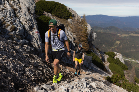 Zwei Wanderer, die Sportkleidung und Rucksäcke tragen, klettern an einem sonnigen Tag auf einem felsigen Bergpfad. Der Hintergrund zeigt zerklüftete Felsen, spärliche Bäume und entfernte Hügel unter einem klaren Himmel.