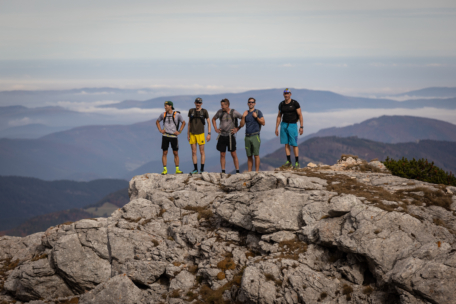 Fünf Wanderer stehen auf einem felsigen Berggipfel und posieren mit Rucksäcken für ein Foto. Der Hintergrund zeigt sanfte Hügel, ferne Berge und einen bewölkten Himmel.