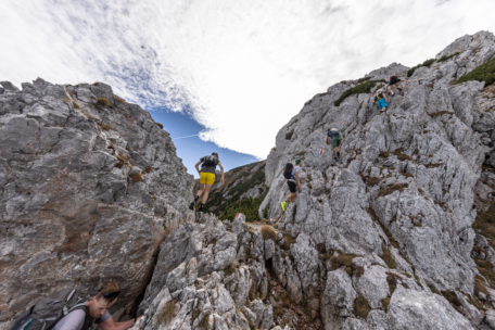 Mehrere Wanderer klettern auf einem steilen, felsigen Bergpfad unter einem teilweise bewölkten Himmel durch zerklüftetes Gelände zwischen zerklüfteten grauen Felsen. Die Szene vermittelt den Eindruck von Abenteuer und Herausforderung in einer dramatischen alpinen Umgebung.