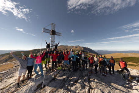 Eine Gruppe von Wanderern posiert freudig mit erhobenen Armen auf einem Berggipfel in der Nähe eines großen Metallkreuzes, unter einem klaren blauen Himmel mit vereinzelten Wolken und einer malerischen Landschaft im Hintergrund.