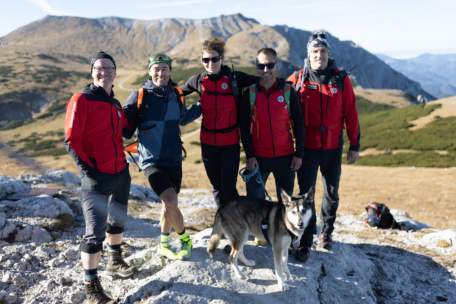 Fünf Wanderer in roten und schwarzen Jacken stehen gemeinsam auf einem felsigen Bergpfad, lächeln in die Kamera und haben einen Hund vor sich. Im Hintergrund sind Berge und grasbewachsene Hänge unter einem klaren Himmel zu sehen.