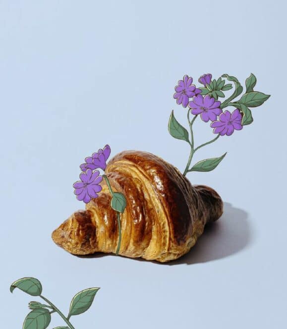Ein goldbraunes Croissant auf hellem Hintergrund, aus dem lilafarbene Blüten und grüne Blätter zu wachsen scheinen, verbindet echte Lebensmittel mit skurriler digitaler Kunst.