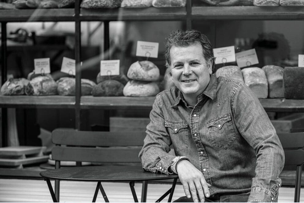 Ein Mann in einem Jeanshemd sitzt an einem Tisch im Freien vor dem Schaufenster einer Bäckerei, in der verschiedene Brotsorten ausgestellt sind. Er lächelt und schaut in die Kamera. Das Bild ist in schwarz-weiß gehalten.