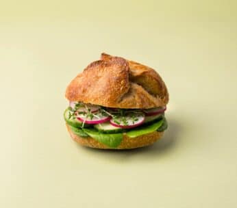 Ein Sandwich mit einem knusprigen Brötchen, gefüllt mit Blattspinat, geschnittenen Radieschen und frischen Sprossen, vor einem schlichten hellgrünen Hintergrund.