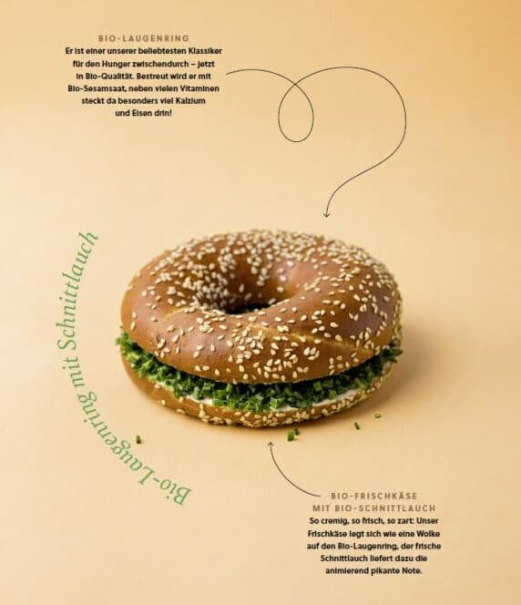 Ein halbierter Sesambrötchen, gefüllt mit Frischkäse und frischem Schnittlauch, ist auf einem hellgelben Hintergrund abgebildet, umgeben von einem deutschen Text, der die Bio-Zutaten beschreibt.