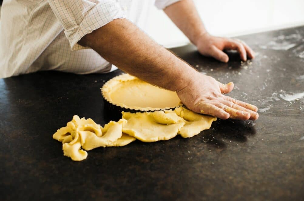 Eine Person drückt den Teig auf einer dunklen Arbeitsfläche in eine Tarteform, wobei der überschüssige Teig über den Rand hängt und beide Hände die Kruste formen.