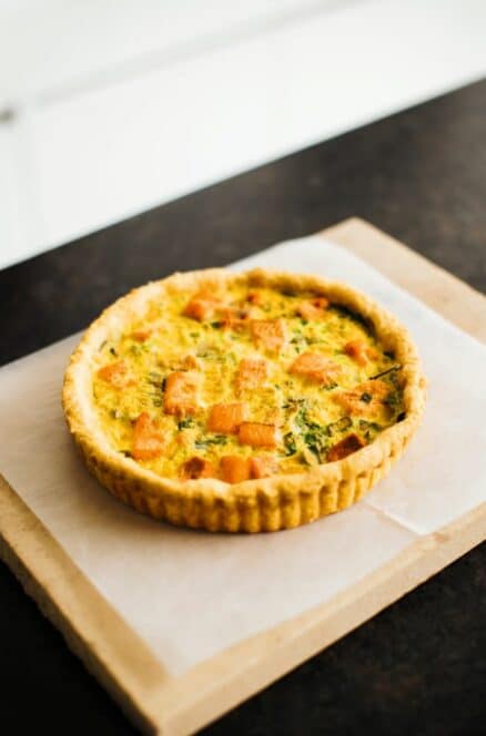 Eine frisch gebackene Quiche mit einer goldenen Kruste und sichtbaren Gemüsestücken liegt auf Pergamentpapier auf einem Schneidebrett aus Stein.