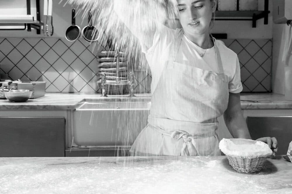 Eine Frau in einer Schürze streut Mehl über einen Küchentisch, während sie einen Korb mit Teig hält und sich auf das Backen vorbereitet. Der Küchenhintergrund zeigt gestapelte Teller und Kochutensilien. Das Bild ist in schwarz-weiß gehalten.
