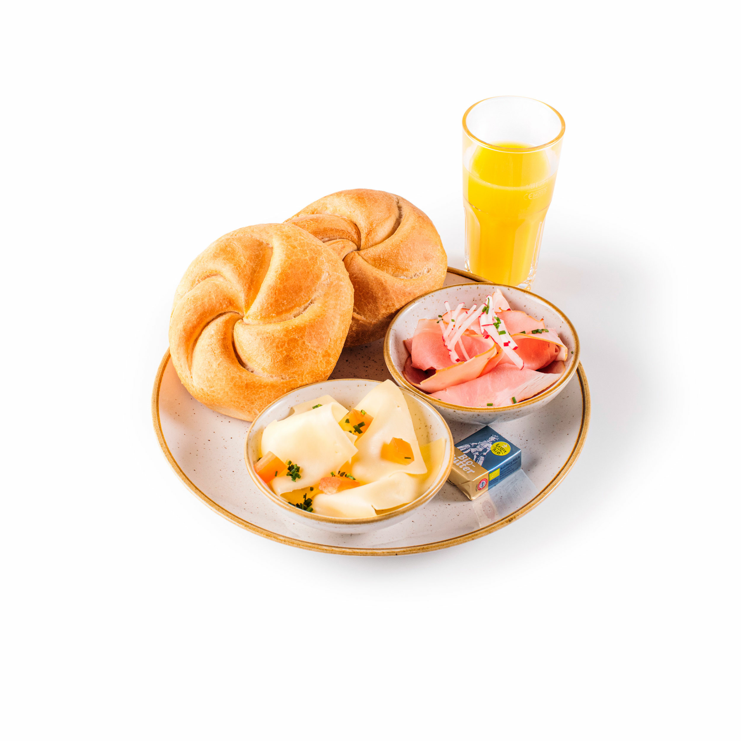 Ein Frühstücksteller mit zwei Brötchen, einer Schüssel mit Käsescheiben, einer Schüssel mit Aufschnitt und Zwiebeln, einem eingewickelten Butterfladen und einem Glas Orangensaft auf weißem Hintergrund.