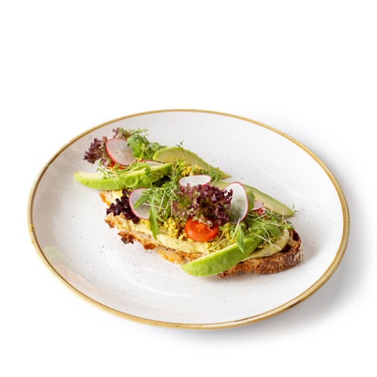 Offenes Sandwich auf rustikalem Brot, belegt mit Avocadoscheiben, gemischtem Blattgemüse, Kirschtomaten, Radieschenscheiben und frischen Kräutern, serviert auf einem weißen Teller mit Goldrand.