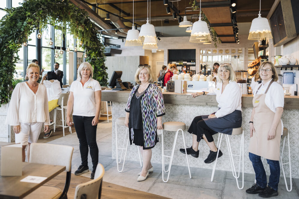 Fünf Frauen posieren und lächeln in einem modernen, hellen Café mit stilvollem Dekor; einige stehen an Tischen, andere sitzen an einem Bartresen, mit Pflanzen und Hängelampen im Hintergrund.