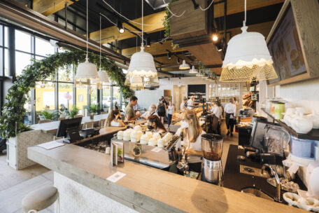 Helles, modernes Café mit großen Fenstern, Hängelampen und Grünpflanzen. Die Baristas bereiten hinter dem Tresen Getränke zu, während die Kunden bestellen und warten. Die Leute sitzen an Tischen und genießen ihre Speisen und Getränke.