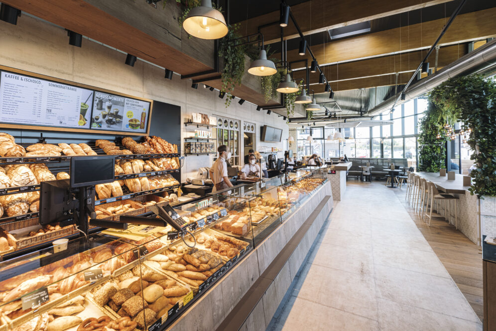Moderne Bäckerei mit Glasvitrinen, in denen eine Vielzahl von Brotsorten und Gebäck angeboten wird. Das Personal bedient die Kunden hinter dem Tresen. Der Raum ist hell, mit hölzernen Akzenten, Hängelampen und Grün um den Sitzbereich herum.