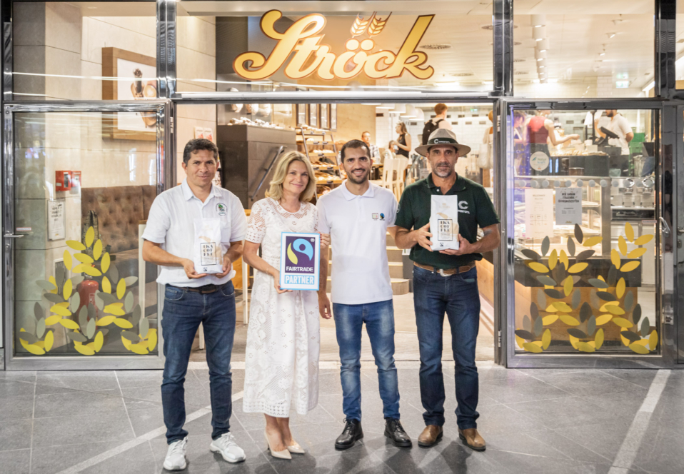 Vier Personen stehen vor einer Bäckerei namens Ströck und halten Kaffeetüten und ein Fairtrade-Partner-Schild in der Hand. Hinter ihnen sind der gläserne Eingang und das Innere der Bäckerei zu sehen.