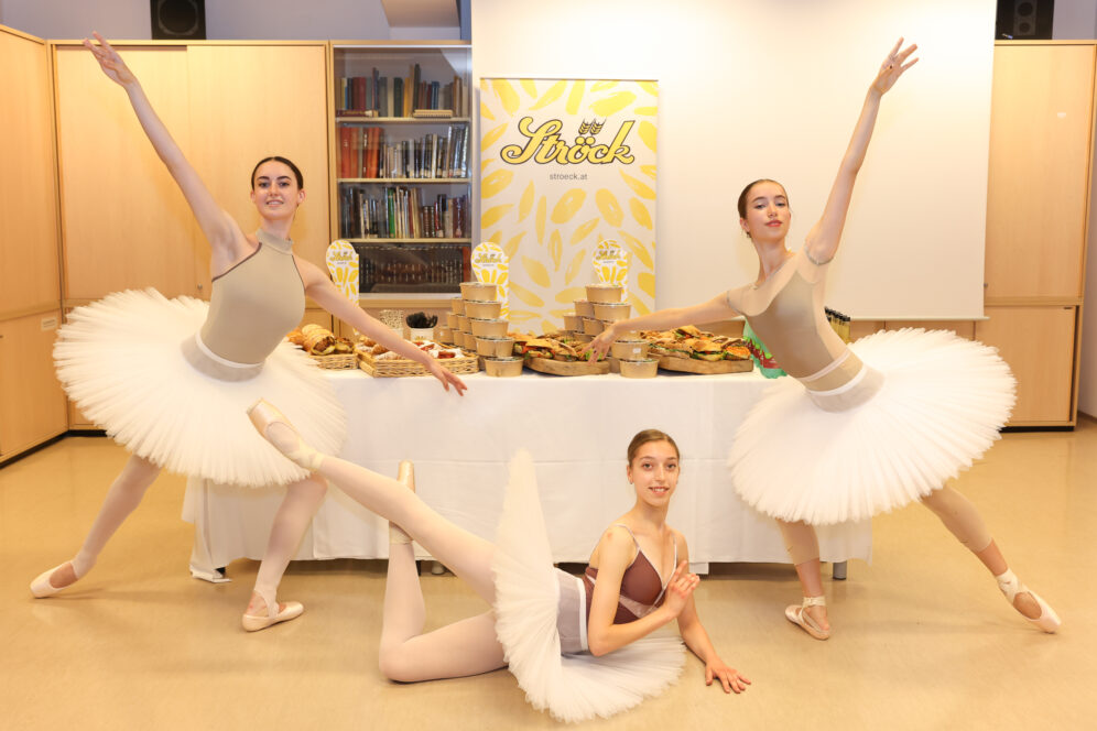Drei Balletttänzerinnen in weißen Tutus posieren anmutig vor einem mit Gebäck und Brot gefüllten Tisch, mit einem dekorativen Ströck-Banner und Bücherregalen im Hintergrund.