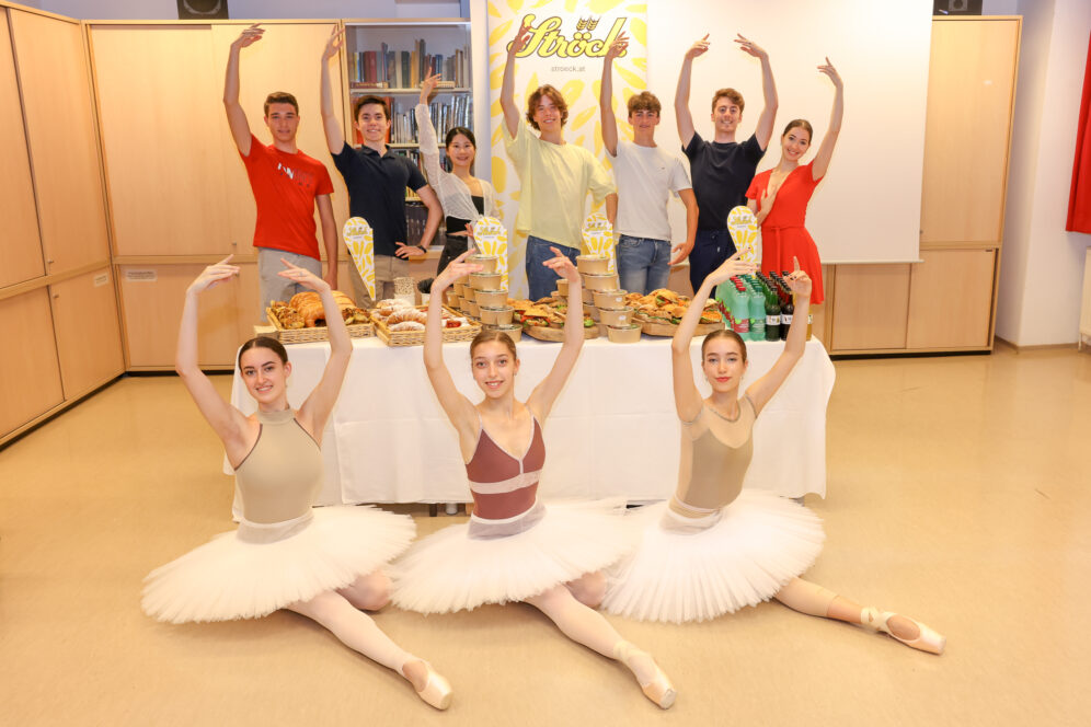 Vier Balletttänzerinnen in weißen Tutus posieren vor einem Tisch mit Essen und Trophäen. Hinter ihnen stehen sechs lächelnde Menschen, die einen Arm heben und die Pose der Tänzerinnen nachahmen. Die Szene wirkt festlich und feierlich.