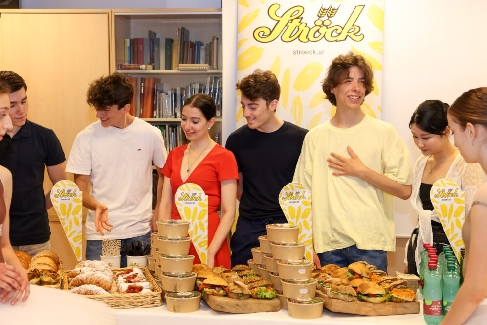 Eine Gruppe junger Leute steht hinter einem Tisch mit belegten Brötchen, Gebäck und Getränken, lächelt und unterhält sich an einer Lebensmittelauslage der Marke Ströck. Im Hintergrund sind Bücherregale zu sehen.