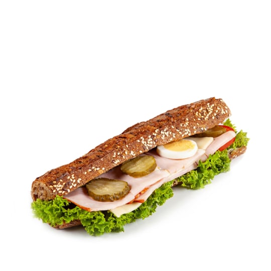 Ein Vollkorn-Baguette-Sandwich, gefüllt mit Salat, einem in Scheiben geschnittenen hartgekochten Ei, Essiggurken, Tomaten und Wurstwaren, isoliert auf einem weißen Hintergrund.