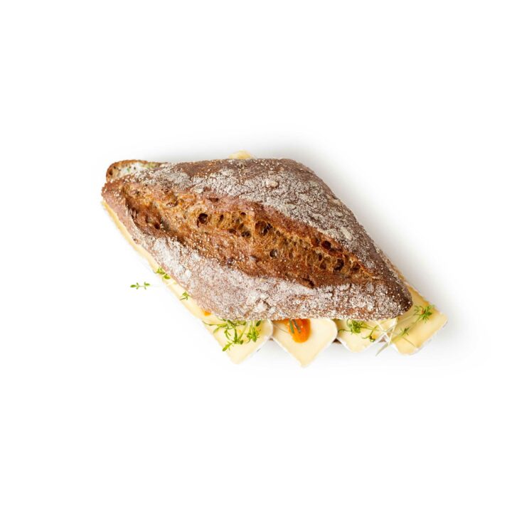 Ein rustikales Vollkornsandwich mit sichtbaren Körnern, gefüllt mit Käsescheiben, Kräutern und Orangengarnitur, auf weißem Hintergrund.