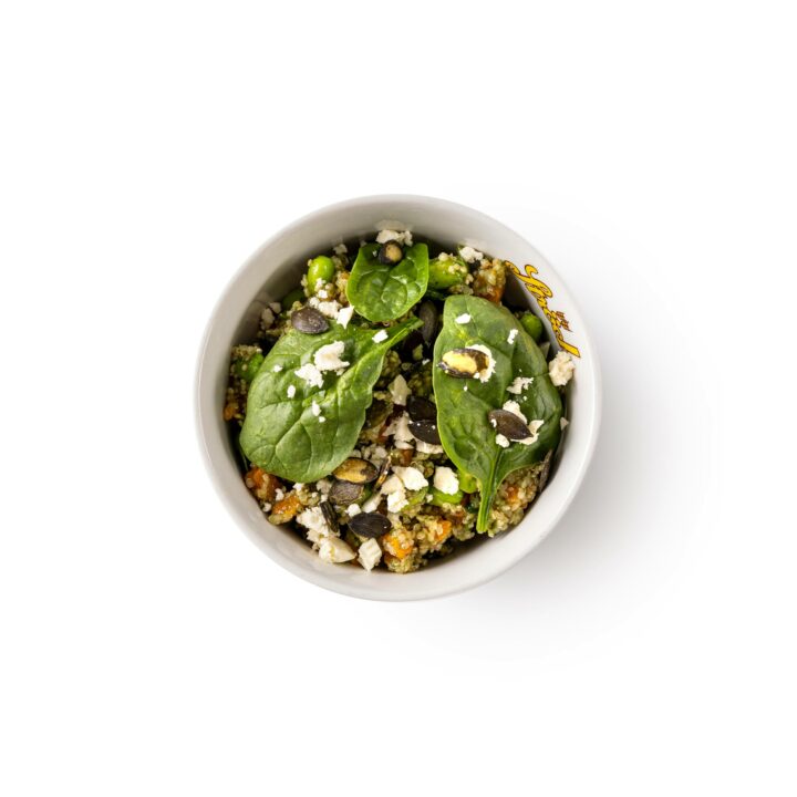 Eine weiße Schüssel mit einem Salat aus Blattspinat, Quinoa, Fetakäse, Kürbiskernen und Gemüsestückchen, von oben gesehen auf weißem Hintergrund.