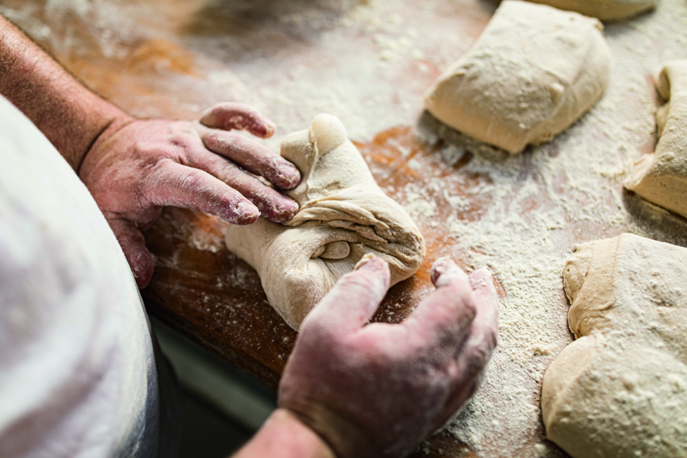 Eine Person mit mehlbestäubten Händen faltet Teigstücke auf einer hölzernen Oberfläche, umgeben von anderen Teigstücken, und bereitet so Brot oder Gebäck zu.