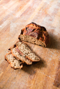 Ein rustikaler Laib handwerklich hergestellten Brotes, teilweise aufgeschnitten, ruht auf einer Holzfläche. Drei Scheiben sind vor dem verbleibenden ungeschnittenen Teil angeordnet. Das Brot hat eine knusprige, goldbraune Kruste und ein luftiges Inneres.