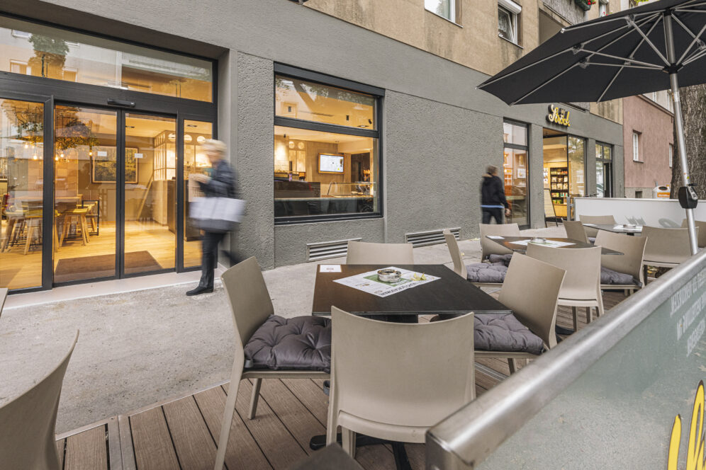 Ein modernes Straßencafé mit leeren Tischen und Stühlen steht neben einem grauen Gebäude; zwei verschwommene Personen gehen am Eingang vorbei, und große Fenster geben den Blick auf ein gemütliches Interieur frei.