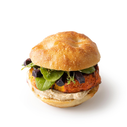 Ein Sandwich mit einem runden, knusprigen Brötchen, gefüllt mit Hummus, einer Gemüsepastete, frischem Blattspinat und gehackten schwarzen Oliven, vor einem schlichten weißen Hintergrund.