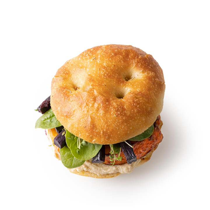 Ein Sandwich auf Focaccia-Brot, gefüllt mit Grünzeug, einer Frikadelle, lila Gemüsescheiben und einem cremigen Aufstrich, fotografiert auf weißem Hintergrund.