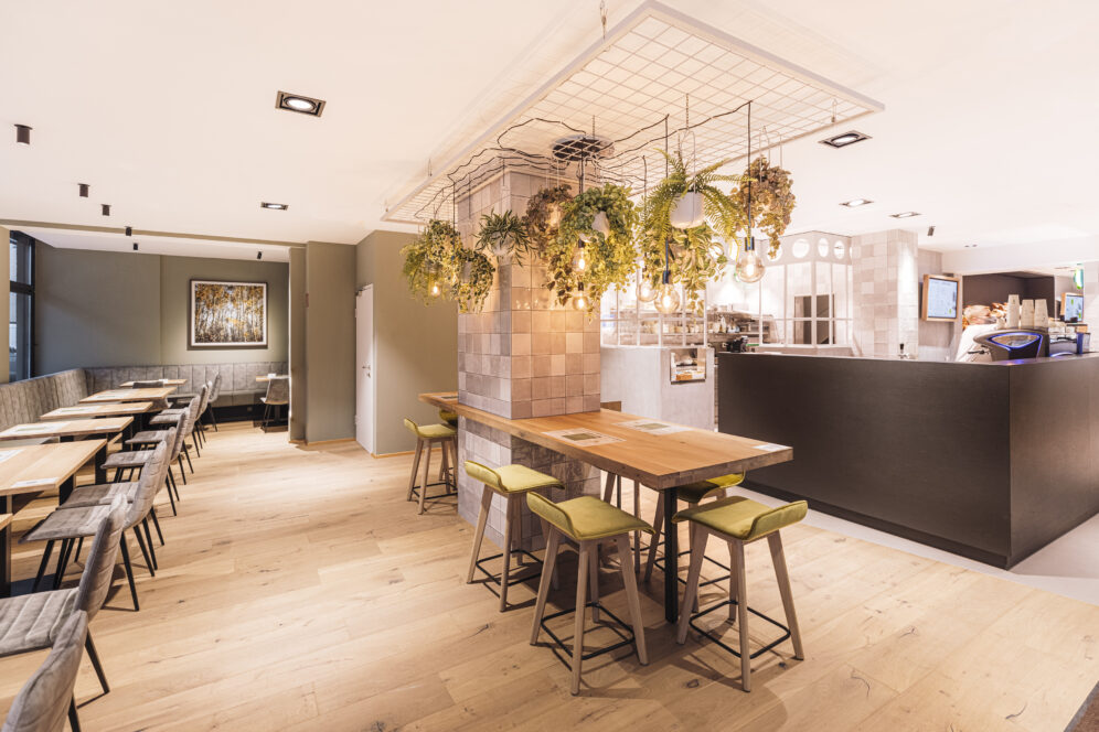 Helles, modernes Café-Interieur mit hellen Holzböden, Tischen und grün gepolsterten Hockern. Pflanzen hängen über einem zentralen Stehtisch. An der Wand stehen Sitzgelegenheiten und im Hintergrund ist die offene Küche zu sehen.
