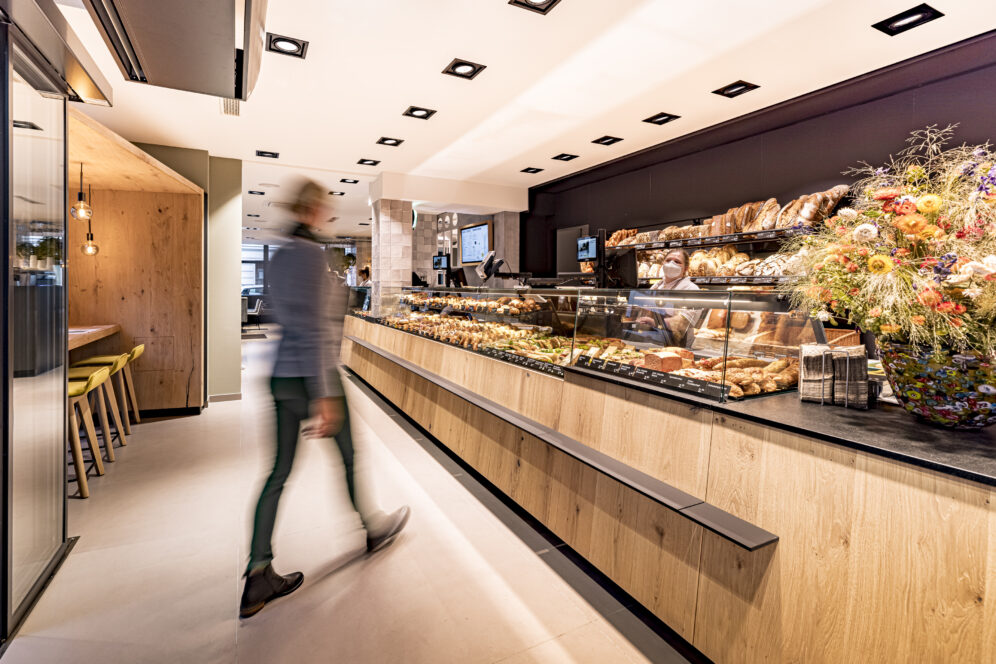 Eine Person geht an einer modernen Bäckertheke vorbei, die mit verschiedenen Brotsorten und Gebäck gefüllt ist. Der helle, saubere Raum ist mit hölzernen Akzenten, Barhockern und einem großen Strauß bunter Blumen auf der rechten Seite ausgestattet.