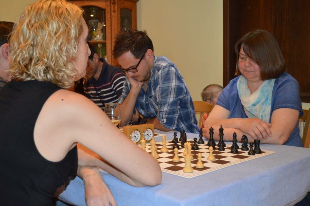 Vier Erwachsene sitzen um einen Tisch und spielen Schach, mit Blick auf ein Schachbrett und eine Uhr. Eine Frau mit einem blauen Schal beobachtet aufmerksam das Brett. Ein kleines Kind sitzt im Hintergrund. Die Atmosphäre ist konzentriert und locker.